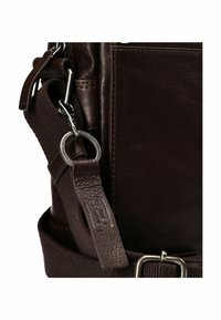 Primer plano de un bolso de cuero marrón que presenta una correa texturizada, herrajes metálicos y una etiqueta distintiva de cuero con la marca.