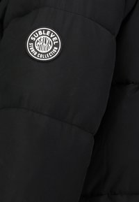 Schwarze Pufferjacke mit einem runden schwarz-weißem Logo-Patch, auf dem "SUBLEVEL STUDIO COLLECTION" steht. Glatte Stofftextur.
