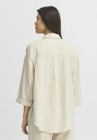 Camicia di lino beige chiaro con vestibilità morbida, maniche lunghe rimboccate, piega centrale sul retro e orlo curvo, caratterizzata da un design del colletto classico.