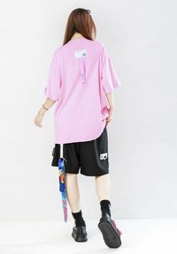 Camiseta rosa de gran tamaño con mangas cortas, combinada con pantalones cortos negros con bolsillos y etiquetas de colores, usada con calzado negro.