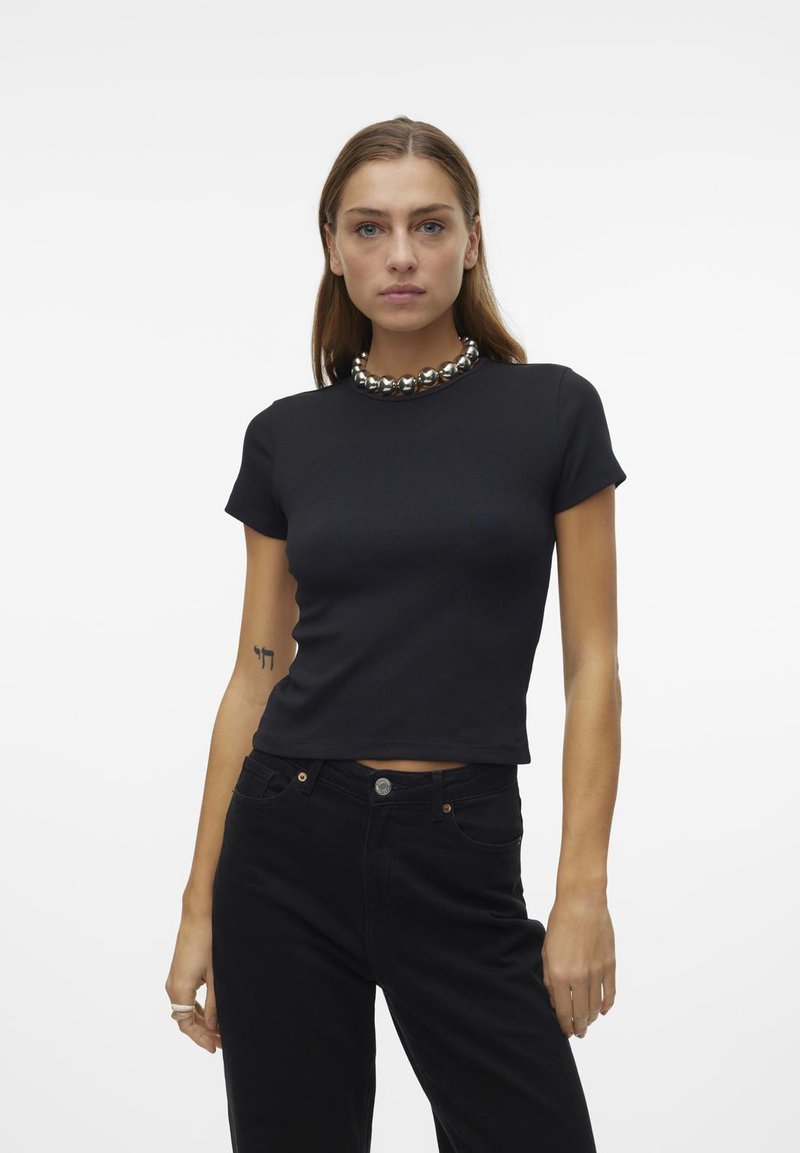 Svart ribbet kortermet topp med rund hals og kort passform, kombinert med høytlivde svarte jeans. Sølvperle-halskjede som aksent.