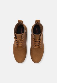 Botas de cuero marrón hasta el tobillo con puntas redondeadas, cordones a juego, paneles laterales texturizados y una pequeña etiqueta de la marca en el costado.