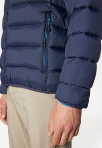 Mornarska puffer jakna s kvadratnim dizajnom, ima džep na zip, elastične manžete i kontrastnu unutrašnjost svijetloplave boje.