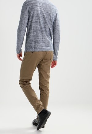 Mand iført grå sweater, brune bukser og sorte sko går væk på en ensfarvet lys baggrund.