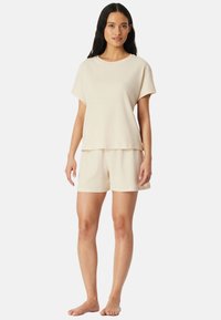 Frau steht barfuß und trägt ein hellbeiges Kurzarmoberteil sowie passende weite Shorts vor einem schlichten weißen Hintergrund.