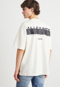 Kremfarget oversized t-skjorte med en svart panel som har teksten "ALLSAINTS" og "LONDON" på baksiden. Korte ermer, avslappet passform, laget av bomull.