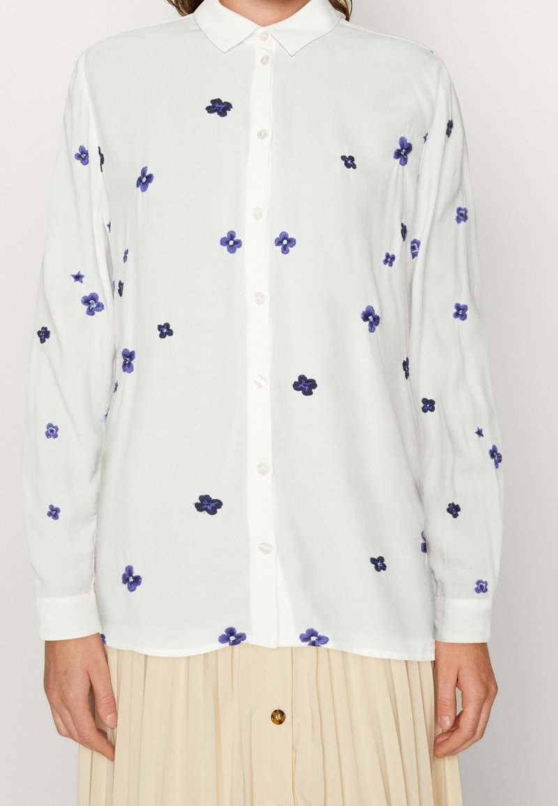 Chemise blanche à manches longues avec petites broderies florales violettes et bleu foncé, portée avec une jupe plissée beige.