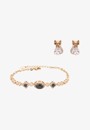 Kultainen rannekoru mustien ja kirkkaiden jalokivien kanssa kullanväristen kettukorvakorujen vieressä, jotka pitävät pisaranmuotoisia kristalleja.