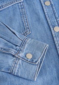 Tessuto in denim azzurro chiaro con cuciture visibili, caratterizzato da un polsino con bottoni e una piccola tasca a pattina con bottoni argentati. Texture liscia.