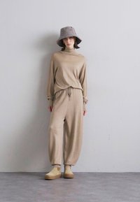 Beige knit outfit with long sleeves and elastic waistband; relaxed-fit pants. Accessorized with a patterned bucket hat and beige slip-on shoes.