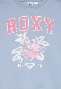 Γαλάζια μπλούζα με έντονο λογότυπο "ROXY" σε ροζ χρώμα, με λευκή και ροζ σχεδίαση από λουλούδια τζαμπαϊκών και την ένδειξη "Est. 90" από κάτω.
