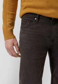 Muž oblečený v horčicovožltom pletenom svetri a tmavo hnedých nohaviciach, s ľavou rukou uvoľnenou pri boku na jednoduchom svetlom pozadí.