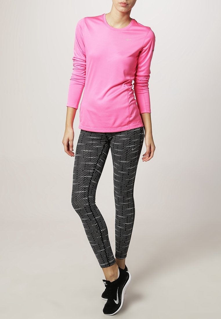 Femme portant un haut à manches longues rose vif, des leggings noirs et blancs à motifs, et des chaussures de sport noires, debout devant un fond uni.