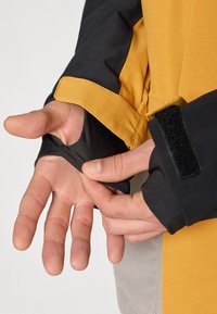 Čierna a žltá vonkajšia bunda s rukávmi na suchý zips; látka vyzerá hladko a odolne; ruka viditeľne upravuje fit rukáva.