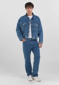 Denim jakna in kavnem modrem, z kontrastnim šivanjem, gumbnimi zaklopkami ter klasičnim krojem in stranskimi žepi.