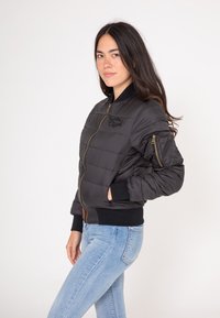 Czarna, ocieplana kurtka bombowca z zapięciem na zamek, ściągaczami przy mankietach i logo na klatce piersiowej. Noszona z jasnoniebieskimi dżinsami.