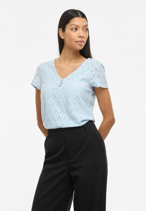 Femme aux longs cheveux noirs portant un chemisier bleu clair à manches courtes avec motif ajouré et un pantalon taille haute noir sur fond blanc.