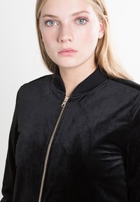 Juodas bomber tipo švarkas, pagamintas iš minkštos, tekstūruotos medžiagos, su auksiniu užtrauktuku, raukšlėtu apykakle ir rankogaliais, pasižymintis elegantišku, minimalistiniu dizainu.