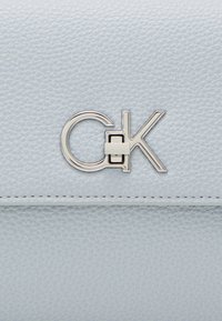 Vaaleansininen teksturoitu keinonahkainen laukku, jossa on hopeinen "CK"-logokiinnike edessä. Ominaisuuksiin kuuluu tikattu reunayksityiskohta ylhäällä.