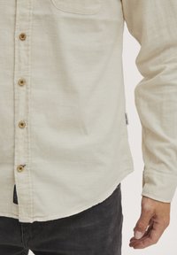 Chemise beige clair avec un design à boutons, dotée de boutons en bois, d'un col et d'une poche poitrine. Le tissu semble doux et texturé.