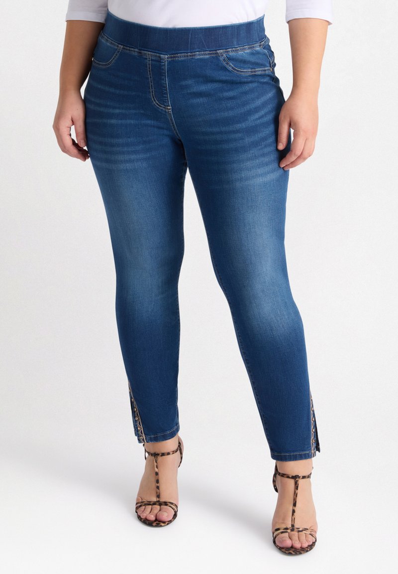 Vaqueros ajustados de denim azul que cuentan con una cinturilla elástica alta, acentos de abertura lateral y una textura suave. Combinados con tacones de estampado animal.