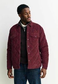 Bordeaux gekleed jasje van corduroy met drukknopen, twee voorzakken met klep en een ronde kraag, gedragen over een donkere trui.