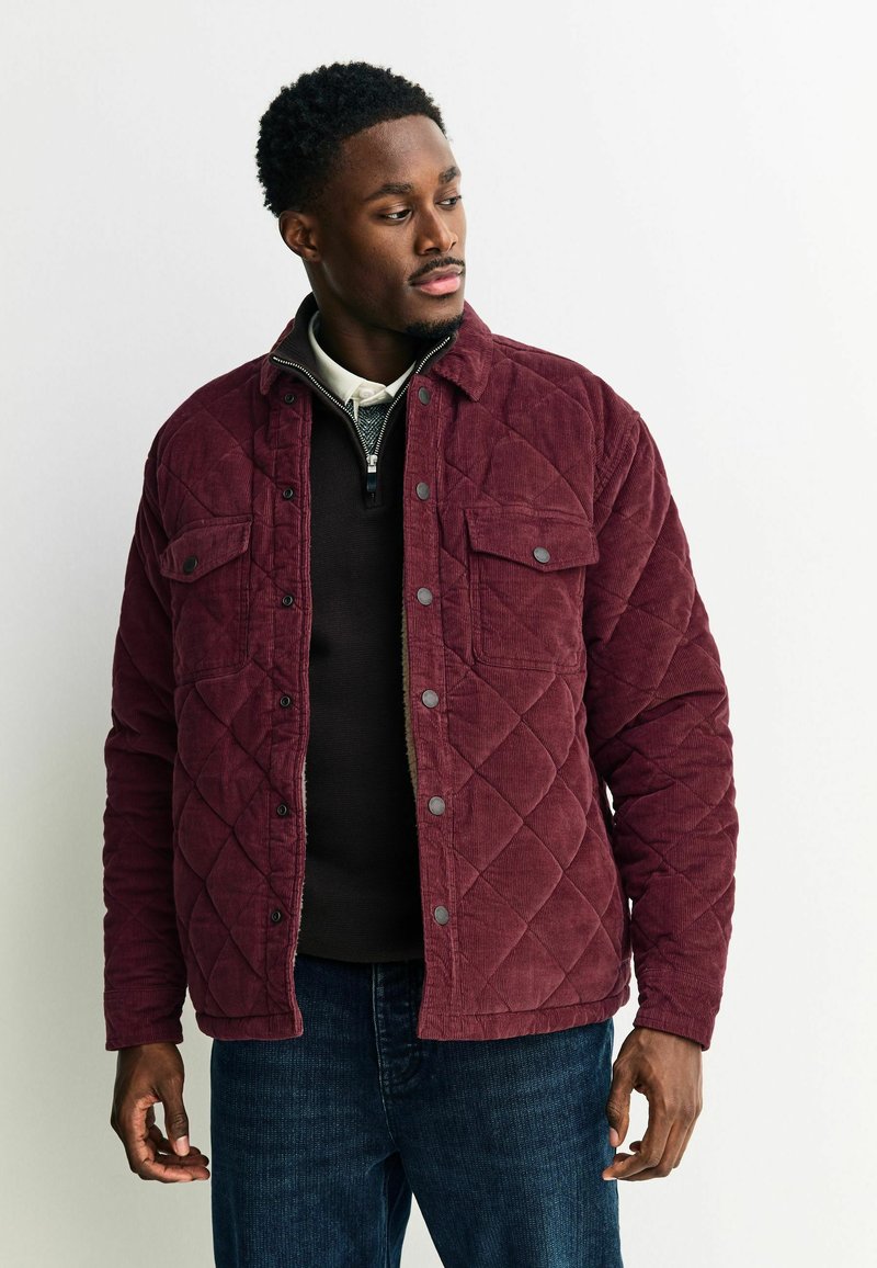 Bordeaux gekleed jasje van corduroy met drukknopen, twee voorzakken met klep en een ronde kraag, gedragen over een donkere trui.