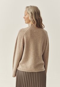 Beige kötött pulóver magas nyakkal, bordás mandzsettákkal és aljjal, mintás szoknyával párosítva. Puha textúra, laza fazon.
