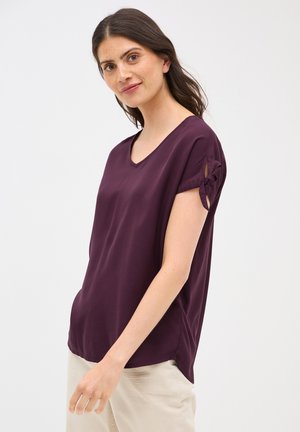 Top a maniche corte e scollo a V in tessuto viola scuro con una consistenza liscia. Presenta un dettaglio annodato alle maniche per un interesse di design aggiuntivo.