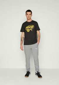 Mees, kellel on seljas must BVB logo T-särk, hallid Puma dressipüksid ja mustad tossud, seisab lihtsa heleda tausta ees.