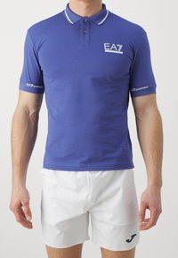 Pikétröja i solid kunglig blå tyg med vit kant på kragen och ärmarna. Har en logotyp på bröstet och varumärkesdetaljer på ärmarna.