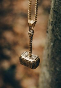 Golden pendant necklace shaped like a hammer, with detailed engravings and a textured handle, hanging against a natural background.