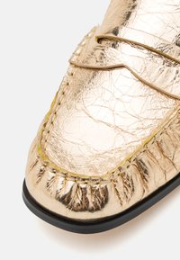Mocassins en cuir doré avec une finition texturée, dotés d'un bord cousu et d'une semelle noire. Comprend une lanière décorative sur le dessus.