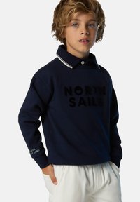 Felpa navy con colletto, caratterizzata da un logo in rilievo "NORTH SAILS", dettagli bianchi e una stampa sulla manica. Indossata con pantaloni bianchi.