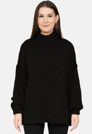 Junge Frau mit glatten braunen Haaren, die goldene Creolen und einen übergroßen schwarzen Strickpullover mit hohem Kragen trägt.