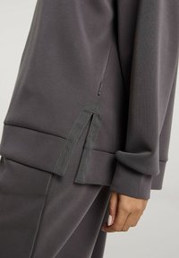 Szara bluza z luźnym krojem i miękką fakturą, z kontrastowym wiązaniem po boku oraz prążkowanymi mankietami.