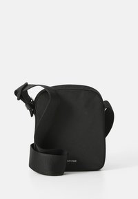 Borsa a tracolla nera in tessuto, con design quadrato, tracolla regolabile e logo del marchio discreto sulla parte anteriore. Texture liscia.