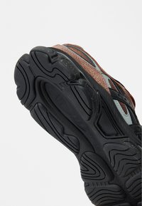 αθλητικό παπούτσι με υφασμένο καφέ και μαύρο πάνω μέρος, λαστιχένια εξωτερική σόλα με περίπλοκα σχέδια πέλματος και διακριτικές μεσοτοιχίες για διαπνοή.