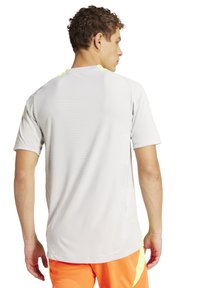 Lichtgrijs sportshirt met korte mouwen, een gestructureerd patroon en felgele accenten op de schouders. Draagt met oranje shorts.
