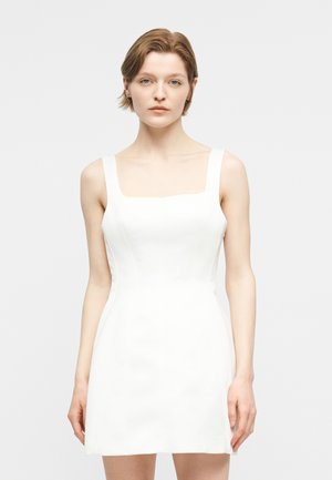 Νεαρή γυναίκα με κοντά καστανά μαλλιά, ντυμένη με αμάνικο λευκό φόρεμα με τετράγωνο ντεκολτέ, στέκεται μπροστά σε απλό ανοιχτό φόντο.