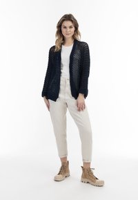 Cardigan in maglia navy con trama aperta, maniche lunghe e bordi a coste, abbinato a una maglietta bianca e pantaloni a tinta crema con gamba affusolata.