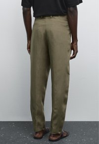 Pantaloni in lino verde oliva con una vestibilità comoda, con fronte piatto, tasca posteriore singola e orli arrotolati, indossati con sandali neri.