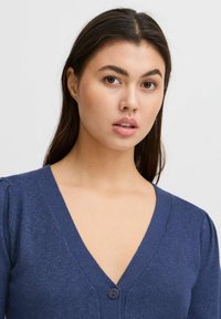 Navy színű, V-nyakú kardigán, texturált kötésből készült, egy gombos záródással és puffos ujjakkal a részletgazdagság érdekében.