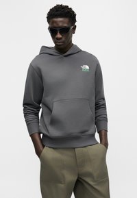 Vyras dėvi tamsiai pilką gobtuvinį megztinį su priekiu kišene, alyvuogių žalių spalvų kelnes, rankas kišenėse ir juodus akinius nuo saulės, žiūrintis tiesiai.