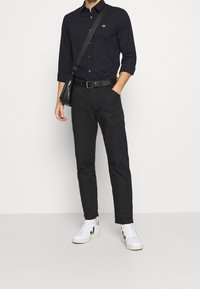 Černá košile s dlouhým rukávem zapínaná na knoflíky, černé kalhoty a bílé tenisky s černými detaily. Černá kabelka přes rameno. Jednobarevné pozadí.