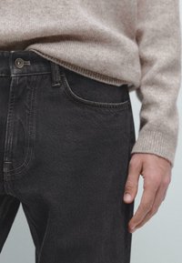 Black denim jeans with contrasting tan stitching, slim fit, mid-rise waist, and side pockets. Worn with a light-colored knit sweater.