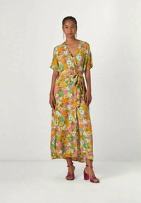Donna che indossa un vestito avvolgente floreale colorato con maniche corte e vita legata, abbinato a sandali con tacco blocco rosa.