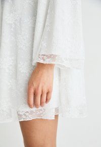 Rochie albă dintr-un material subțire, texturat, cu modele florale, cu mâneci lungi și o detaliere din dantelă pe margine, accentuată de un finisaj neted.