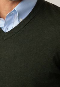 Un pull tricoté vert foncé avec un col en V, porté par-dessus une chemise à col bleu clair. La texture est lisse avec un fin côtelage le long des bords.