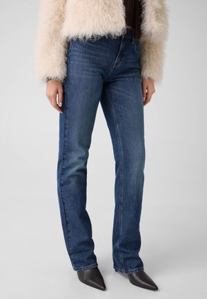 Frau, die gerade geschnittene blaue Jeans, schwarze spitze Schuhe und eine cremefarbene, flauschige Jacke trägt, steht vor einem einfarbigen Hintergrund.